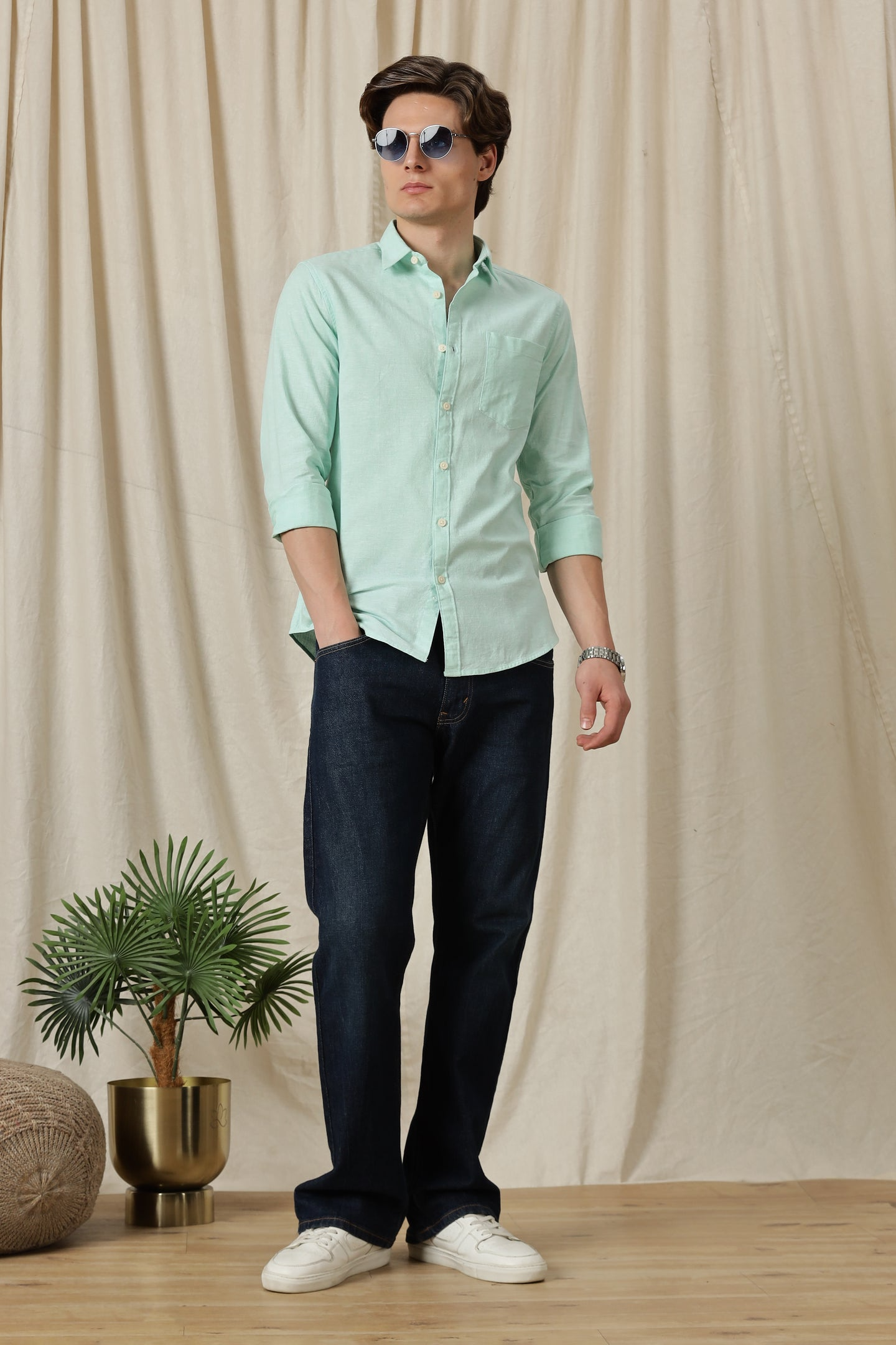 Man wearing a light green shirt and dark pants standing against a beige curtain with a plant in the background.