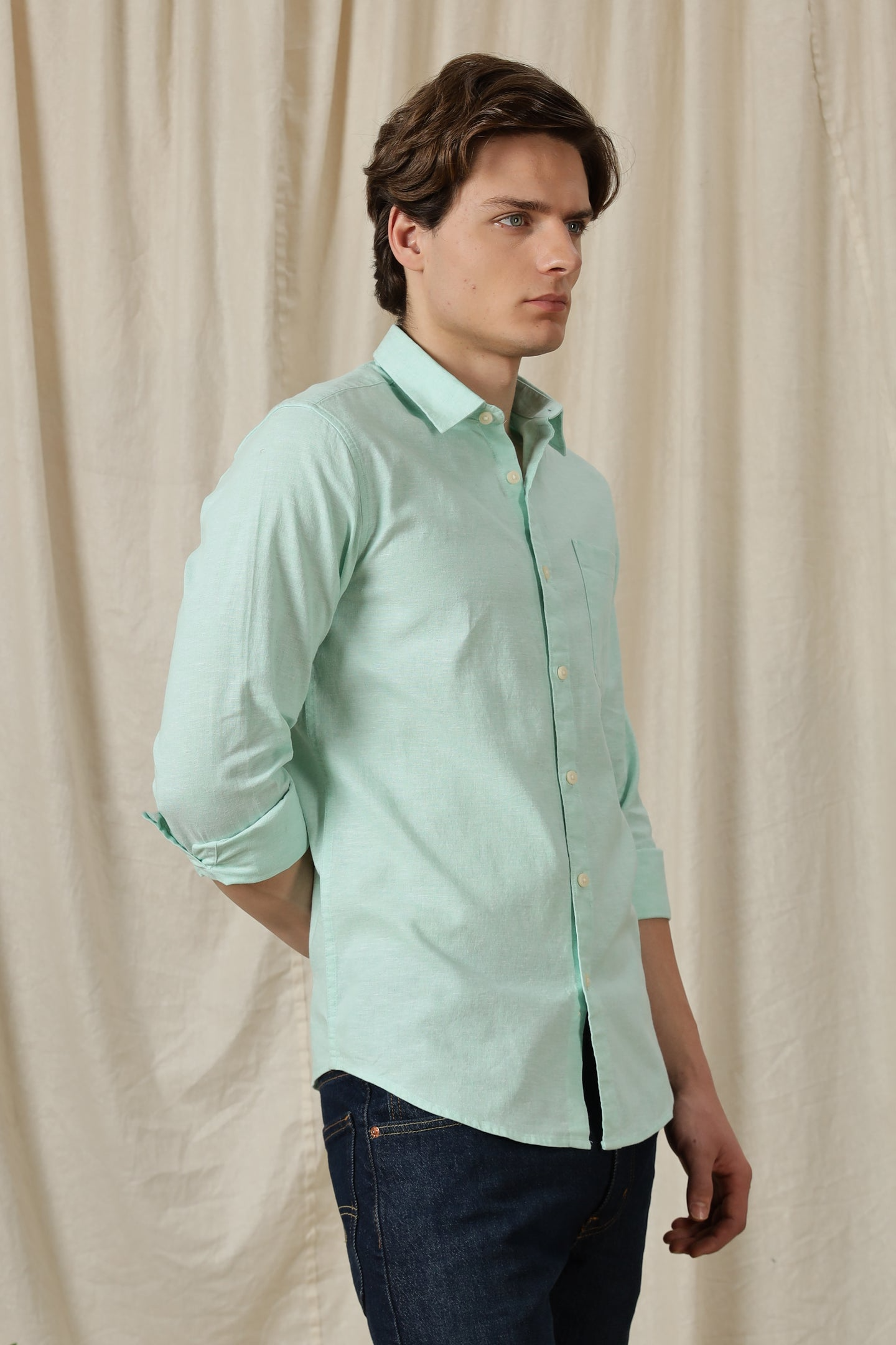 Man wearing a light green shirt and dark jeans against a beige curtain background